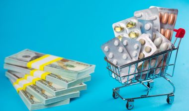 High generic drug prices concept. shopping cart filled red medicinal capsules tablets pills, wad of dollar money, blue background