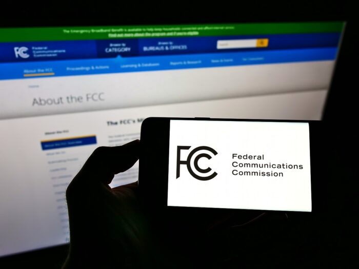 Person holding smartphone with US agency Federal Communications Commission (FCC) seal on screen in front of website.