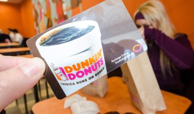 A hand holds up a Dunkin Donuts gift card inside of a Dunkin Donuts franchise store eating area.