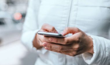 Close up of woman's hands in white jacket holding grey smartphone and typing a message with both hands on street.