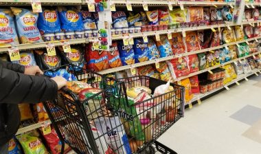 Pushing a cart filled with groceries on the snacks aisle inside a supermarket.