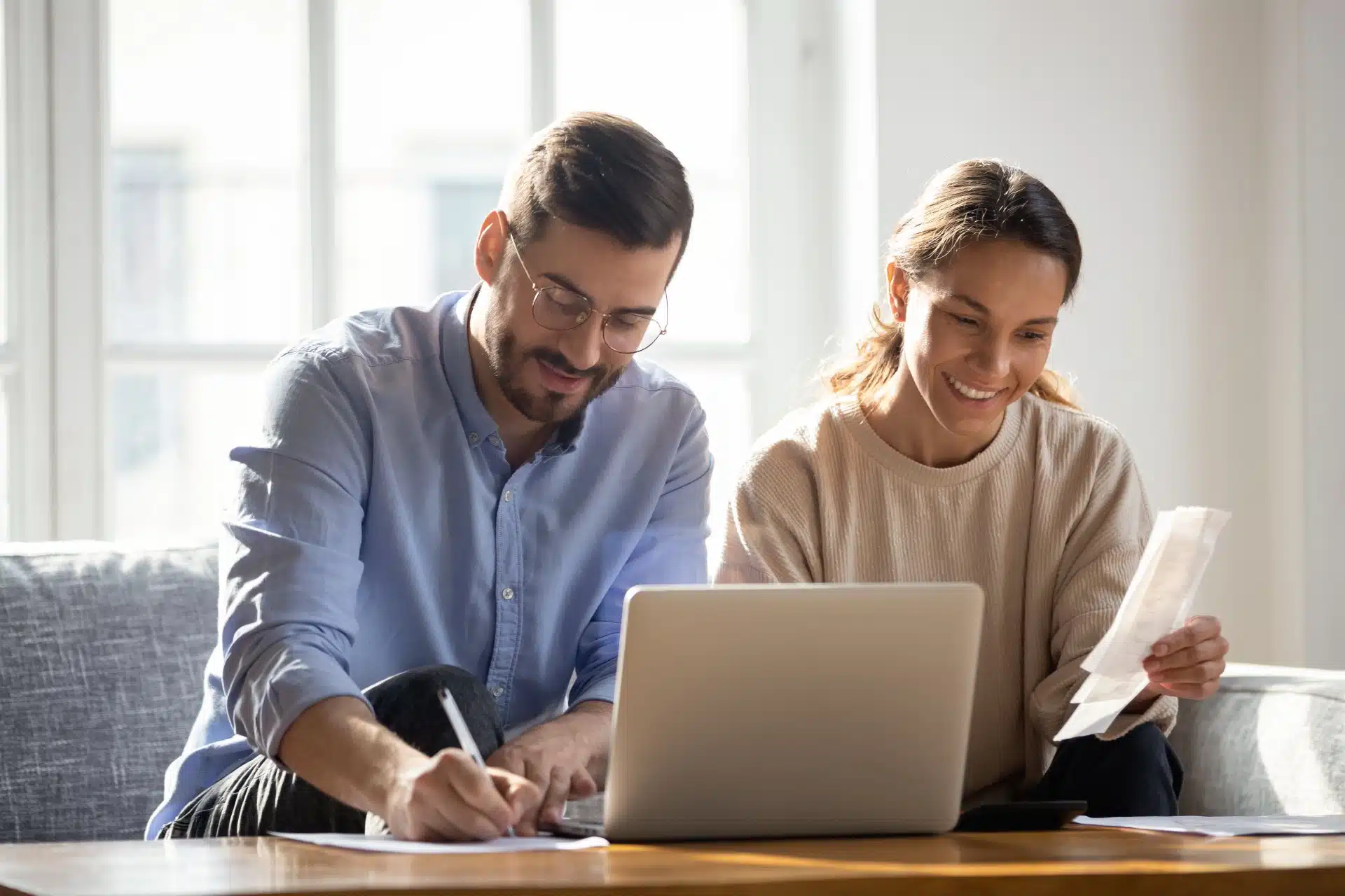 A happy man takes notes next to a laptop while a smiling woman sitting next to him holds up receipts - class action settlement, settlements
