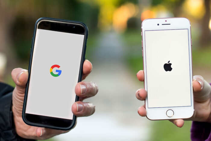 Close up of two peoples hands holding their smartphone. Google logo is displayed on screen. Apple logo is displayed on the other screen.