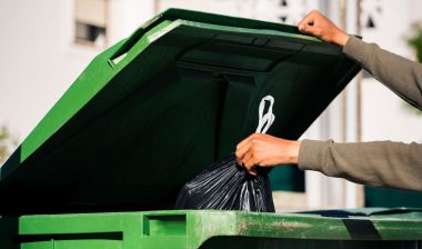Man throwing out black eco-friendly recyclable trash bag in to big plastic green garbage container.