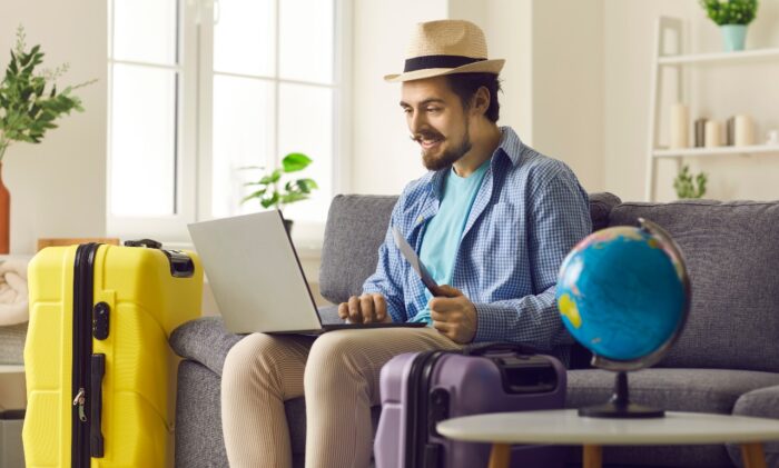 Travel and tour agency online service concept.  Happy smiling man in summer hat holding ticket and passport using laptop digital interface for hotel booking or reservation from home on vacation