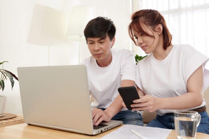 A man types on a laptop while a woman holds up her phone nearby - class action settlements
