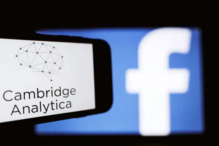 The logo of the strategic communication company Cambridge Analytica is seen on the screen of an iPhone in front of a computer screen showing Facebook logo.
