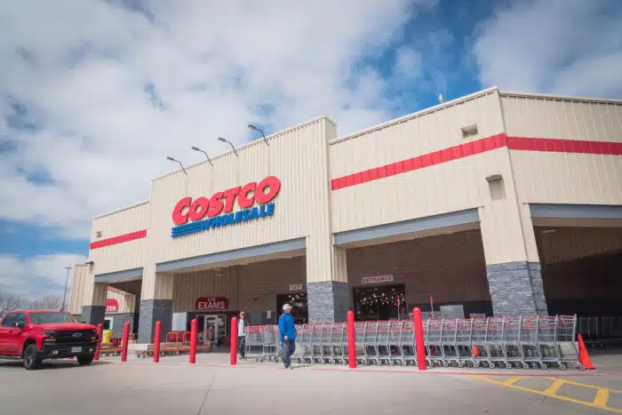 Busy customers walking in and exit the Costco Wholesale store in Lewisville, Texas against a cloudy blue sky.