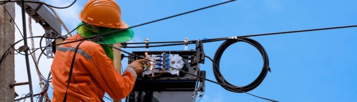 Rear view of technician on wooden ladder checking fiber optic cables in internet splitter box on electric pole against blue sky background