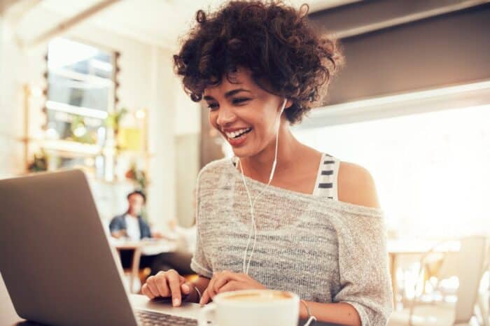 A happy woman wearing earbuds sits in a cafe, working on a laptop - class action lawsuit settlement