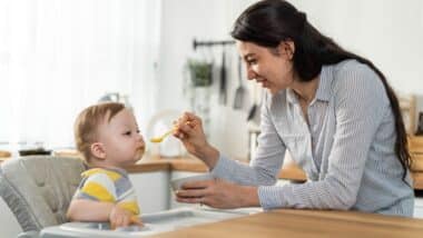 A mother giving her baby food on a spoon inside a kitchen.