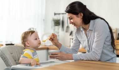 A mother giving her baby food on a spoon inside a kitchen.