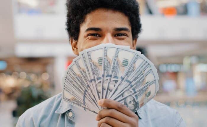 Happy wealthy successful smiling african american business man looking at camera holding fan of money hiding behind dollar banknotes.
