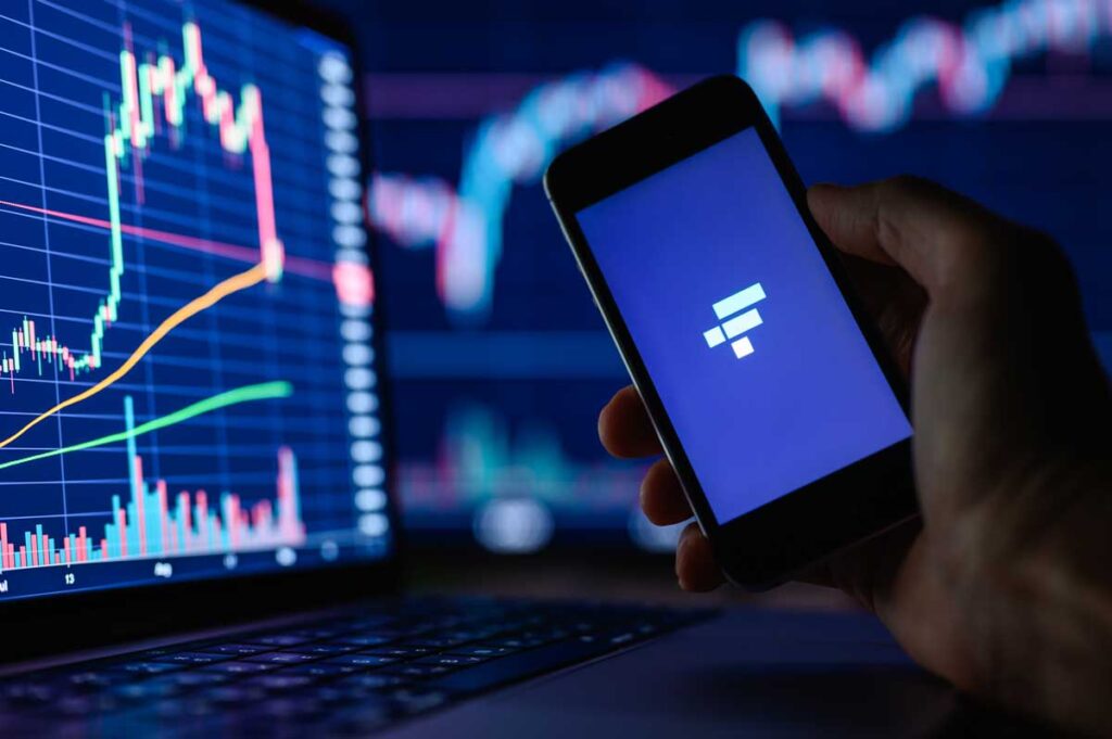 FTX cryptocurrency trading application on a smartphone in hand against the background of monitors with financial charts.