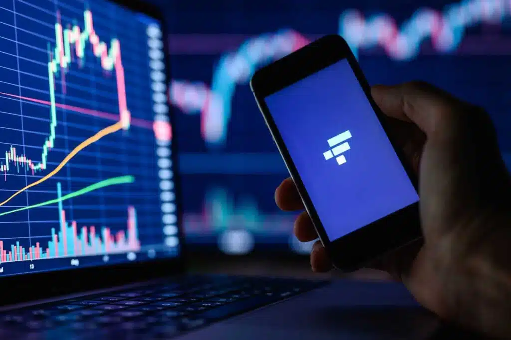 FTX cryptocurrency trading application on a smartphone in hand against the background of monitors with financial charts.