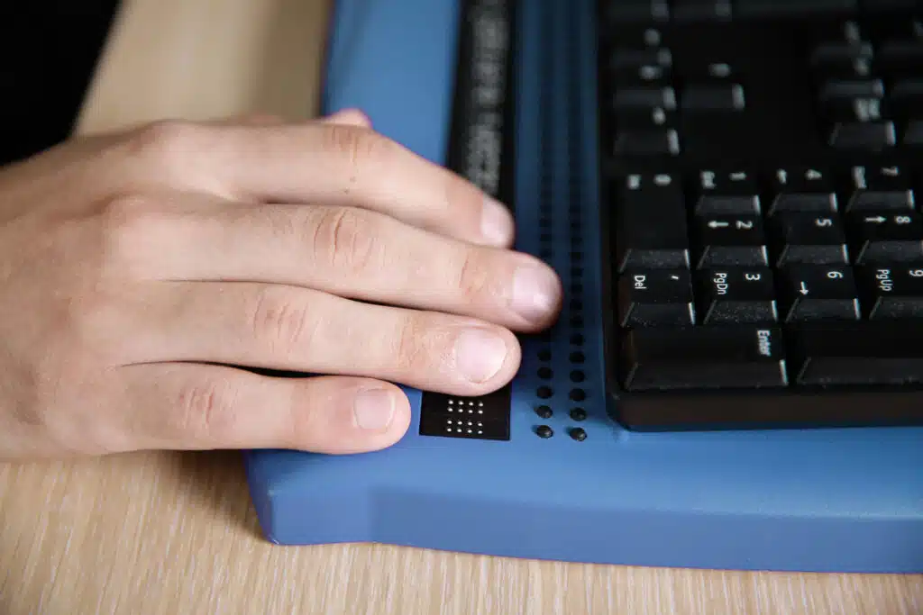 Blind person using computer with braille computer display and a computer keyboard.