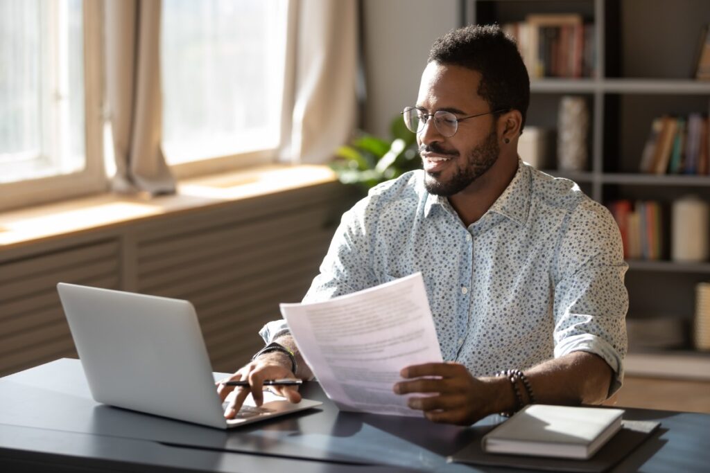 A smiling man in glasses holds papers in his left hand while using a laptop with his left, representing the settlements closing in January 2023.