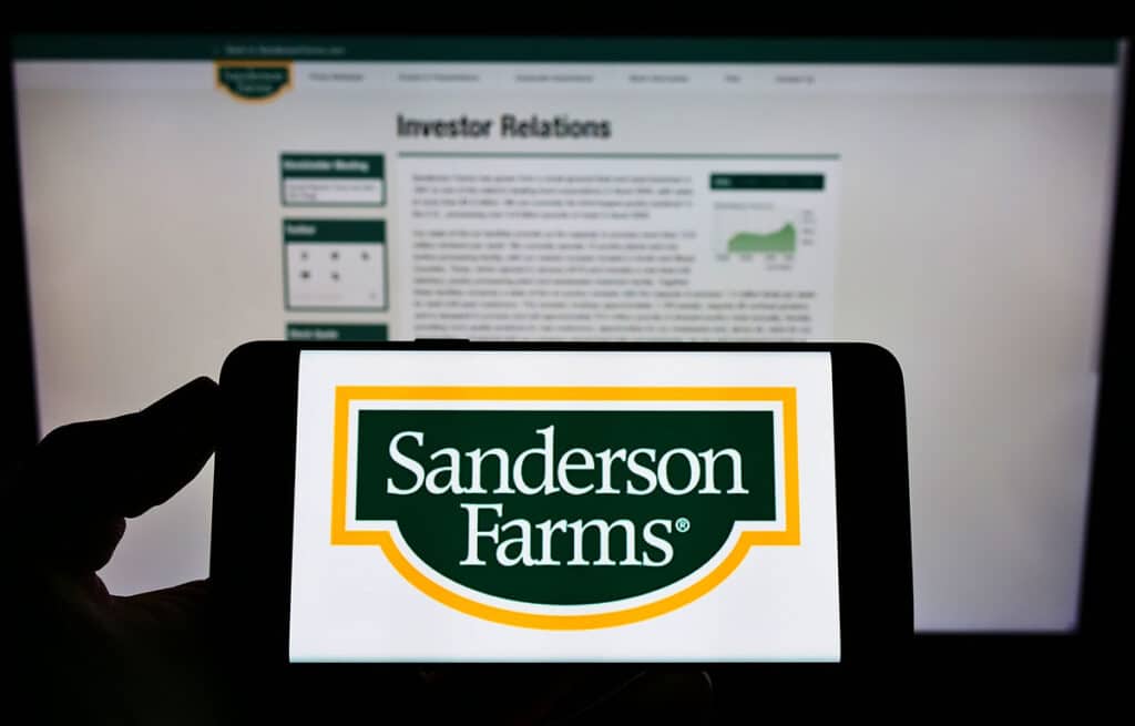 Person holding mobile phone with logo of American poultry company Sanderson Farms Inc. on screen in front of web page.