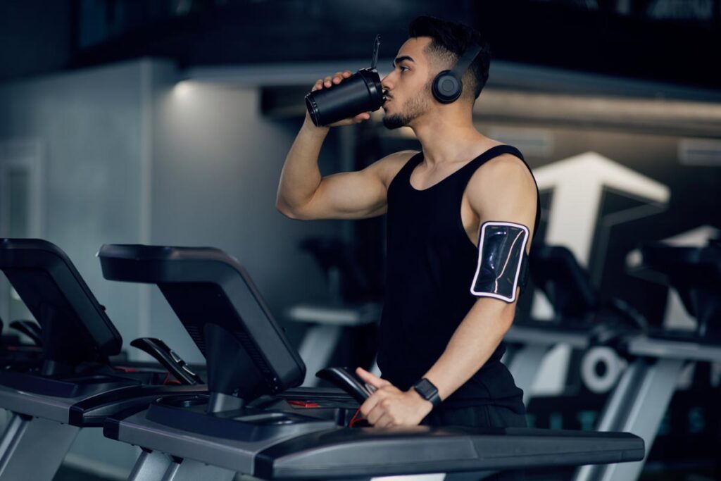 Muscular Arab Guy Drinking Protein Cocktail From Sport Shaker While Training On Treadmill At Gym.