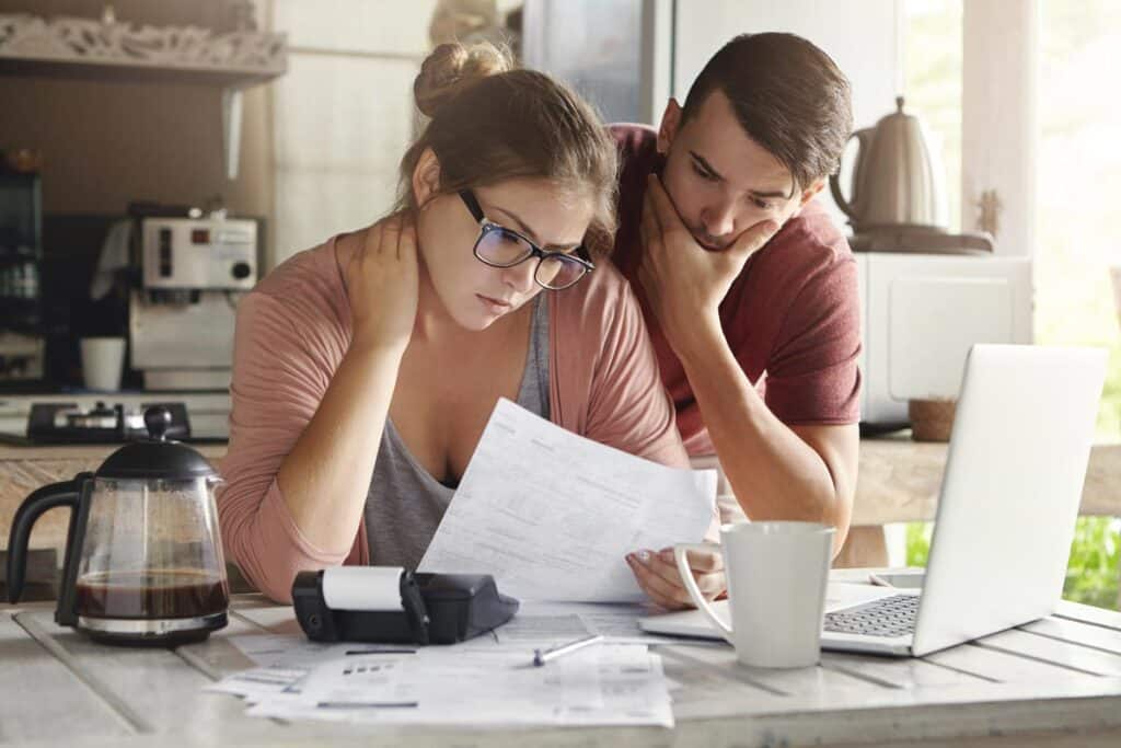Female in glasses and brunette man studying paper form bank while managing domestic budget together in kitchen interior.