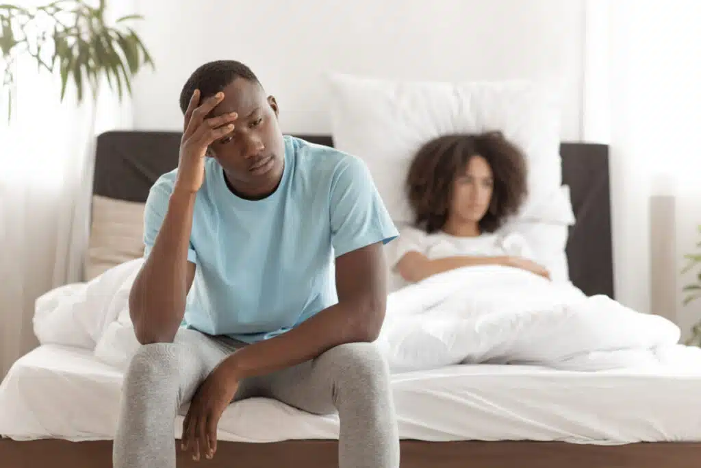 A disappointment man sitting at the edge of a bed with a disappointment woman laying in bed
