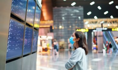 Woman looking at flight information board with mask on representing the Lufthansa settlement.