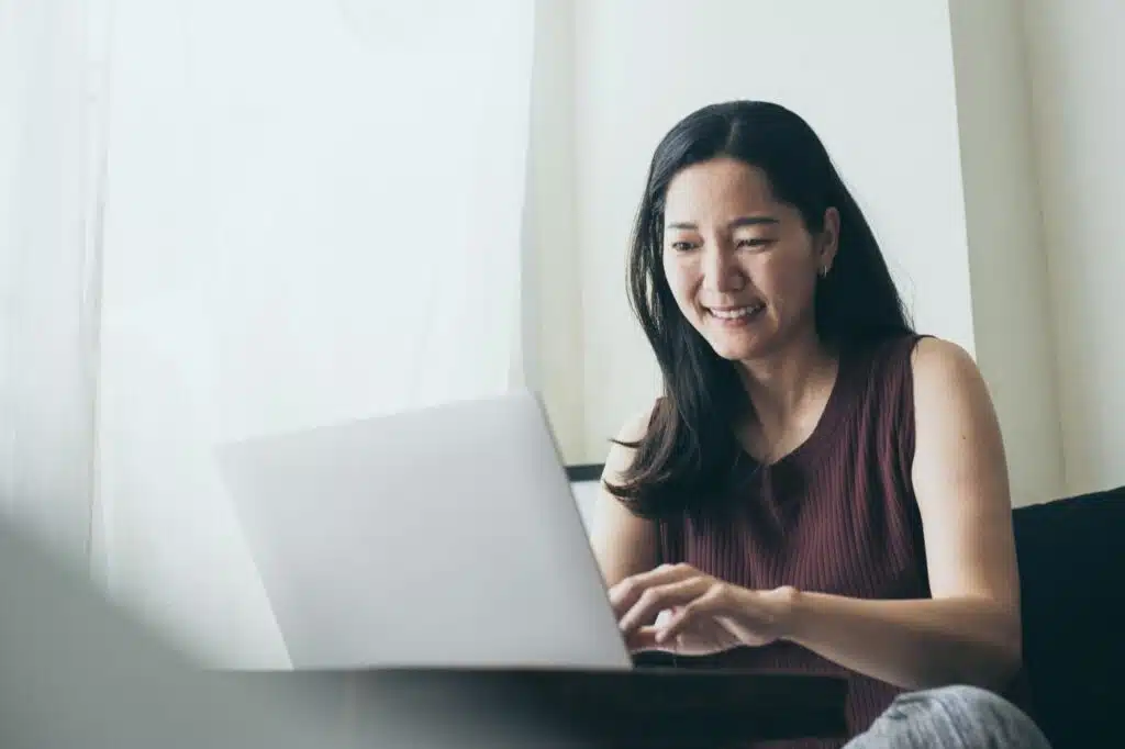 A smiling woman uses a laptop near a window, representing the class action lawsuit settlement closing soon.