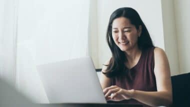 A smiling woman uses a laptop near a window, representing the class action lawsuit settlement closing soon.