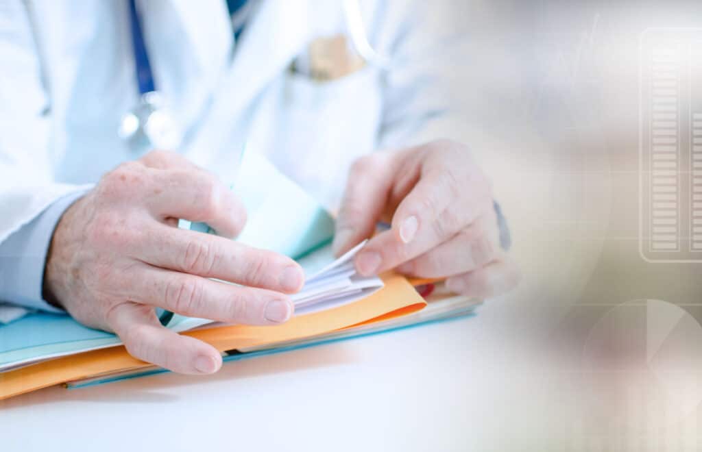 Close up of a doctor looking through a file in a stack of paper on his desk, representing the Medical Review Institute of America (MRIA) data breach class action lawsuit settlement.