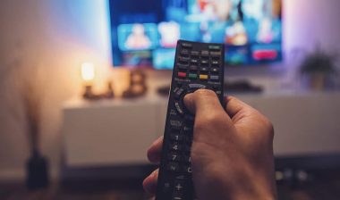 Close up of a mans hands pointing a remote towards a TV, representing FCC cable requirements.