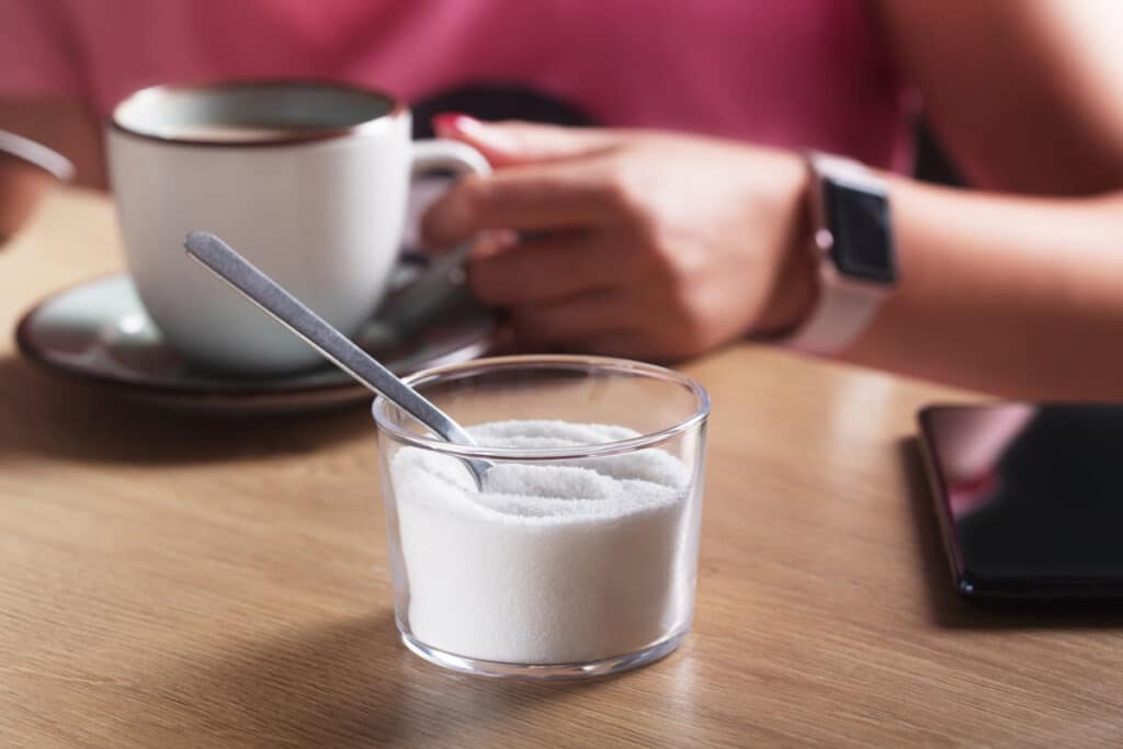 Close up of sweetener and woman drinking coffee in background, representing the WHO's report on aspartame.