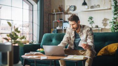 A man sits on a green sofa while holding a paper and using a laptop on his coffee table, representing class action lawsuit settlements closing soon.