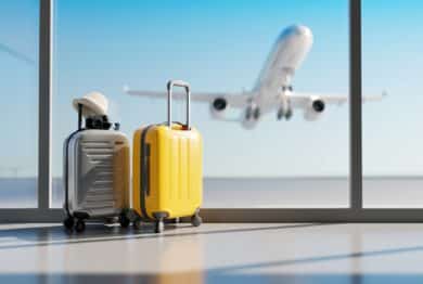 A yellow suitcase and a silver suitcase with a hat and camera on top sit in an airport, with an airplane taking off in the background outside the window, representing the Spirit Airlines settlement.