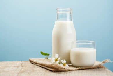 A glass bottle of milk sits next to a short glass of milk, sitting with small white flowers on wooden table against a blue background, representing the Lyons Magnus class action lawsuit settlement.