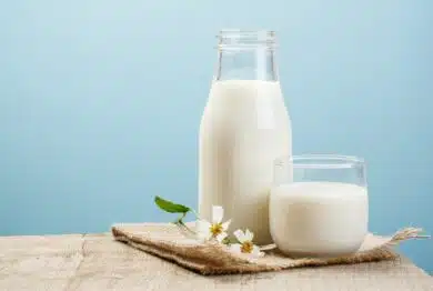 A glass bottle of milk sits next to a short glass of milk, sitting with small white flowers on wooden table against a blue background, representing the Lyons Magnus class action lawsuit settlement.
