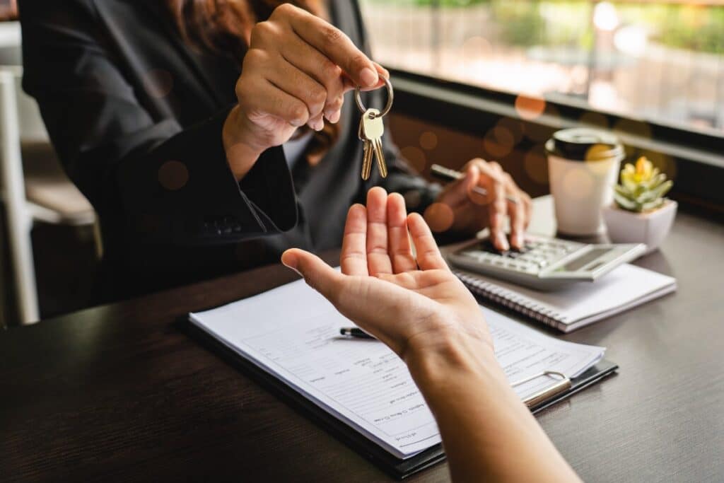 A person in a blazer sits behind a desk, handing a set of keys to an outstretched hand, representing the Re/Max and Anywhere settlement.