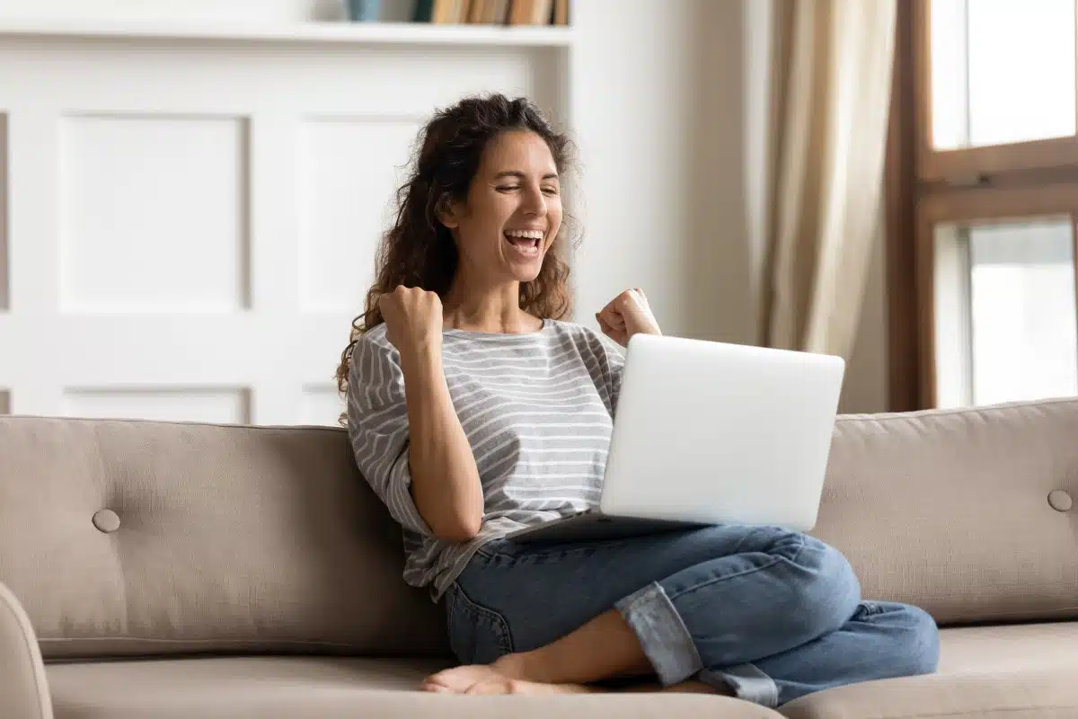 A smiling woman cheers while sitting on her soft with her laptop perched on her legs, representing class action settlements closing soon.