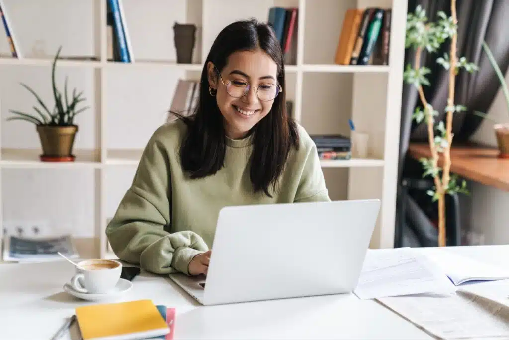 A young woman in glasses sits at a white desk, working on a laptop surrounded by papers and a cup of coffee, representing settlements closing soon.