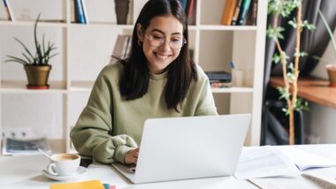 A young woman in glasses sits at a white desk, working on a laptop surrounded by papers and a cup of coffee, representing settlements closing soon.