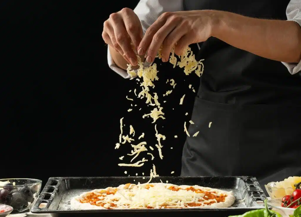 Cook putting shredded cheese on a pizza.