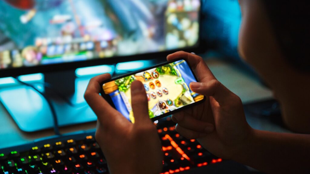teenage gamer boy playing video games on smartphone and computer in dark room wearing headphones and using backlit colorful keyboard