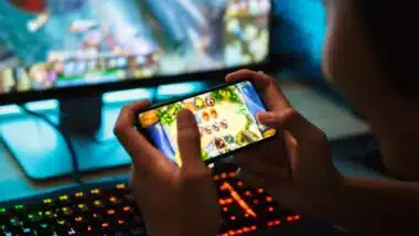 teenage gamer boy playing video games on smartphone and computer in dark room wearing headphones and using backlit colorful keyboard