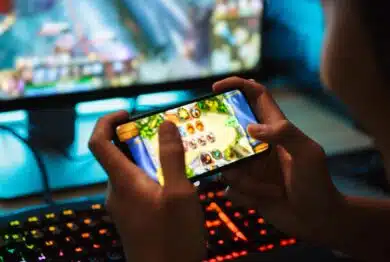 teenage gamer boy playing video games on smartphone and computer in dark room wearing headphones and using backlit colorful keyboard