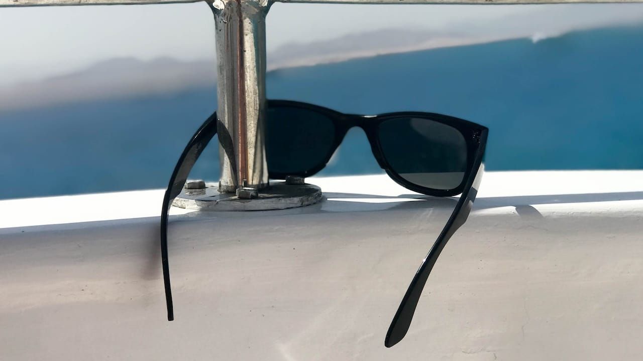 Black plastic sunglasses hang on an iron railing on a yacht, a cruise liner against the blue sea and mountains.