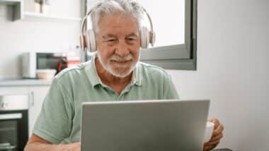 Smiling active modern white haired senior man with headphones sitting at home table using laptop in online connection surfing the net.
