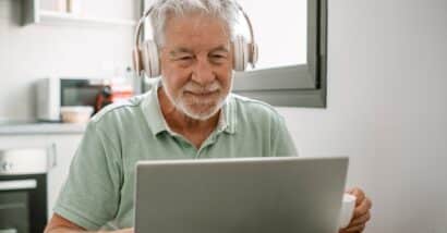Smiling active modern white haired senior man with headphones sitting at home table using laptop in online connection surfing the net.