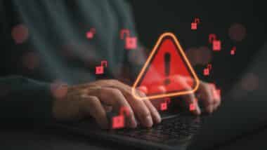 Hands typing on laptop with cybersecurity warning alert and unlocked padlock icons. Data breach risk, hacking attack, ransomware threat, online security vulnerability concept for business protection.