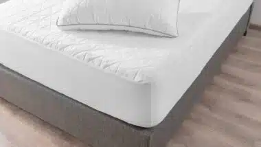 Close-up View of Pristine White Bed, Geometric Quilting on Mattress and Pillow, Contrasting with Soft Gray Upholstered Bed Frame, Comfort, Set Against a Warm Wooden Floor Background.