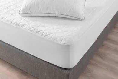 Close-up View of Pristine White Bed, Geometric Quilting on Mattress and Pillow, Contrasting with Soft Gray Upholstered Bed Frame, Comfort, Set Against a Warm Wooden Floor Background.