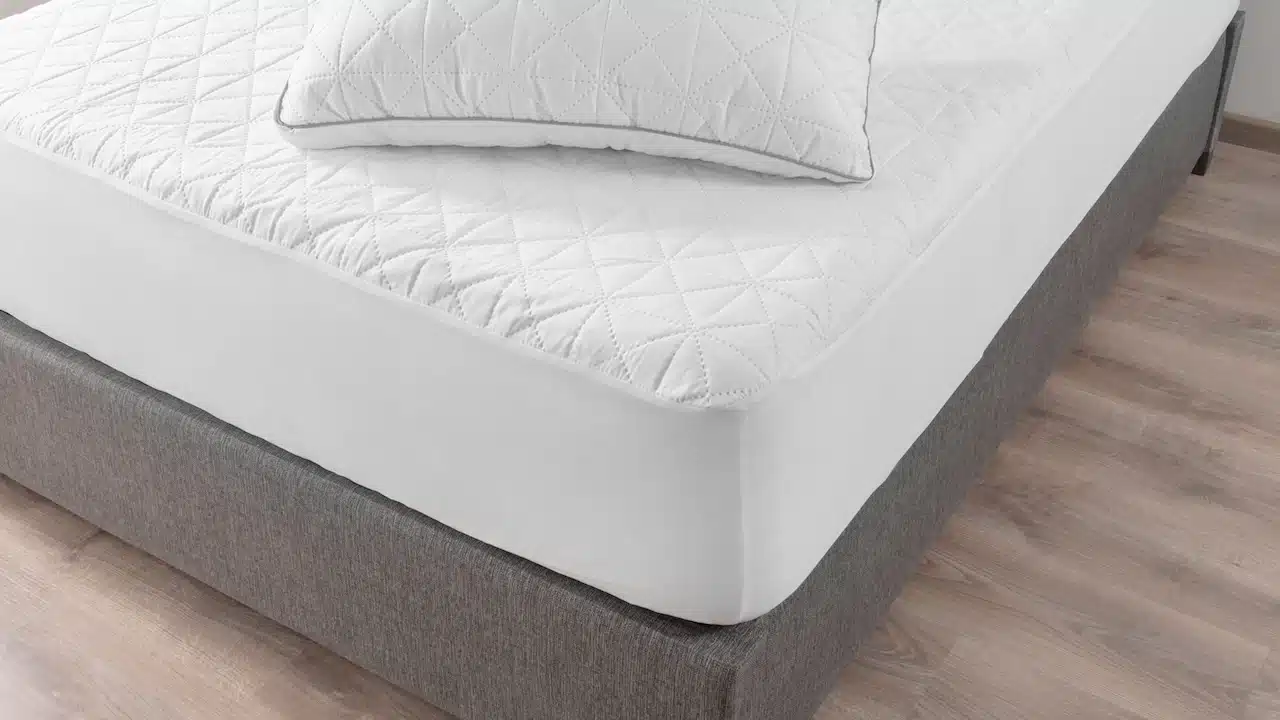Close-up View of Pristine White Bed, Geometric Quilting on Mattress and Pillow, Contrasting with Soft Gray Upholstered Bed Frame, Comfort, Set Against a Warm Wooden Floor Background.